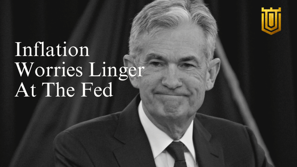 Close-up grayscale portrait of Jerome Powell smiling thoughtfully in a dark suit and striped tie, against a black curtain backdrop. Overlay text: "Inflation Worries Linger At The Fed" in elegant white lettering, with a gold tower-shield logo. Insights from Nicholas Mugalli at World Trade Securities on persistent inflationary pressures and monetary strategy.